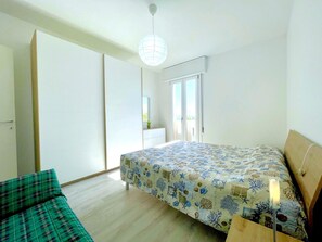 A bedroom with a bed, a green sofa, a sliding door, a balcony, and a hanging light fixture. - Apartment in Porto Santa Margherita (Caorle)