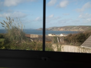 Blick aufs Meer durch ein Fenster, mit einer Wiese, einer Steinmauer und sanften Hügeln im Hintergrund.