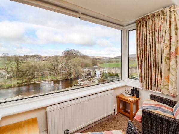 A room with a large window overlooking a river, a radiator, a wicker chair, and a small wooden table. - Riverside, Grassington (Grassington)