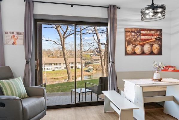 A room with a large sliding glass door, a white table with a vase, a gray sofa, and a poster on the wall. - Clubhouse Suite | Golf & Water View | 85” TV (Branson)