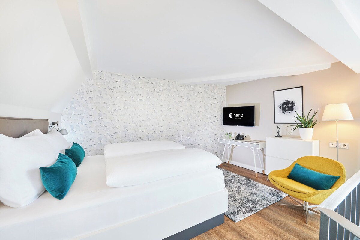 Een moderne woonkamer met een witte bank, blauwgroene kussens, een gele fauteuil, een klein bijzettafeltje, een televisie en een hangende plant.