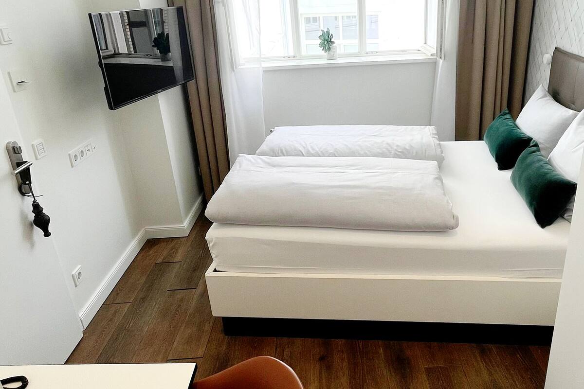 Habitació d'hotel amb un llit, un televisor fixat a la paret, una finestra amb cortines i una petita planta al davant de la finestra.
