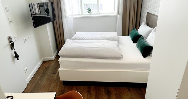 Una habitación de hotel con cama, televisor montado en la pared, ventana con cortinas y una planta pequeña en el alféizar.