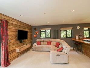 Ein modernes Wohnzimmer mit einer Eckcouch, einem flachbildfernseher, der an einer Holzwand befestigt ist, einer Küchenzeile mit Bar und großen Fenstern.