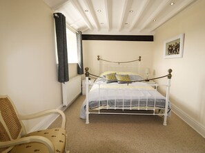 Ein Schlafzimmer mit weißem Metallbett, einem beigefarbenen Teppich, einem Sessel mit gemustertem Kissen und einem Fenster mit dunkler Gardine.