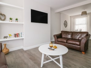 Un salón con un sofá de cuero marrón, una mesa de centro blanca, un televisor de pantalla plana montado en la pared y una ventana con persianas.