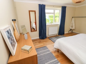 A bedroom with a bed, a wooden bedside table with a book and a vase, a window with curtains, and a radiator.