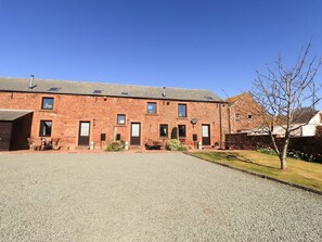 A brick building with multiple windows and doors, a gravel driveway, and a tree in the foreground. - Stable Cottage, Bolton Low Houses (Wigton)