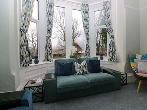 Un canapé vert avec des coussins bleus, une petite table et une chaise dans une pièce dotée de grandes fenêtres et de rideaux à motifs.