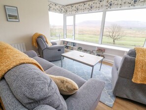 A living room with a sofa, armchair, and a table. There are large windows with a view of the countryside. - Netherscar, Ingleton (Carnforth)