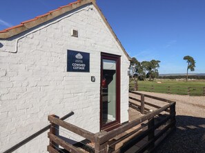 A white cottage with a sign reading 'COWSHED COTTAGE,' a wooden fence, and a clear sky.