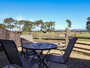 An outdoor seating area with a round table and chairs, a wooden fence, and a view of trees and open land.