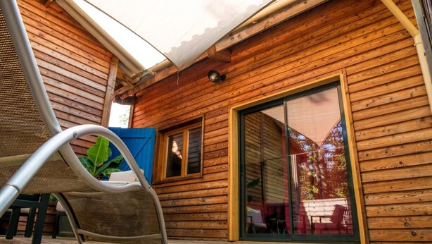 A wooden cabin with a covered patio, a sliding glass door, and a metal chair. - Chalet Jardin Les Fées (Saint-Remèze)