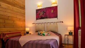 A bedroom with a wooden wall, a bed with a purple bedspread, two bedside lamps, and a wall hanging. - Chalet Jardin Les Fées (Saint-Remèze)