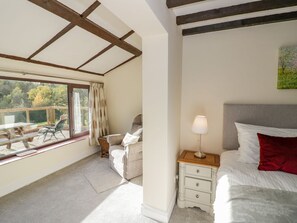 Ein Schlafzimmer mit Bett, Nachttisch mit Lampe, Fenster mit Vorhängen und Blick ins Freie.