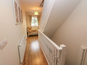 A narrow hallway with a wooden floor, a white radiator, a white railing, and a window with curtains.