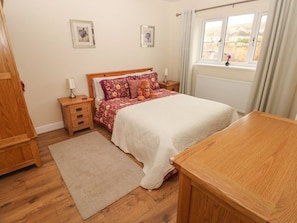 A bedroom with a wooden bed, bedside tables, a nightstand, a window with curtains, and a view of the outside.
