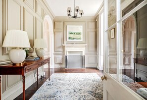 A hallway with a fireplace, a chandelier, and a view of a landscape through a window. - Batheaston House, Batheaston (BATH)