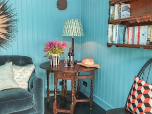A cozy room with a blue wall, a wooden side table with a lamp, a vase of flowers, and a bookshelf. - Foxglove Cottage, Harome (York)