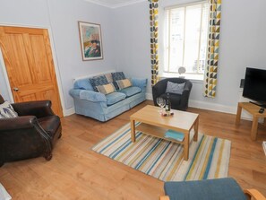 A living room with a wooden floor, a blue sofa, a black armchair, a coffee table, a television, and a window with patterned curtains.