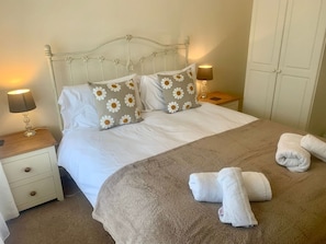 Una cama bien hecha con una colcha blanca y marrón, dos almohadas con estampado de margaritas, dos lámparas de noche y una mesita de noche de madera.