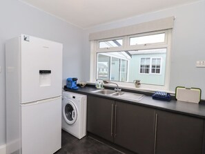 A compact kitchen with a washing machine, a refrigerator, a sink, and a window with a view of a house. - Chatelherault, Ruthwell (Dumfries)