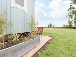 Een blauw gebouw met een raam, een bloembed met planten, een stoel en een grasveld op de achtergrond.