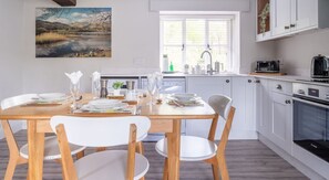 A modern kitchen with white cabinets, a Bosch oven, and a dining area with a wooden table and white chairs. - Grassholme, Hawkshead (Ambleside)
