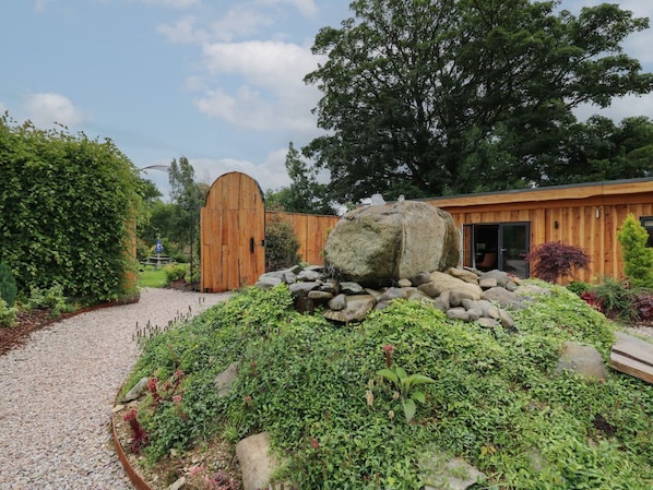 A wooden building with a large rock centerpiece, surrounded by greenery and a gravel path.