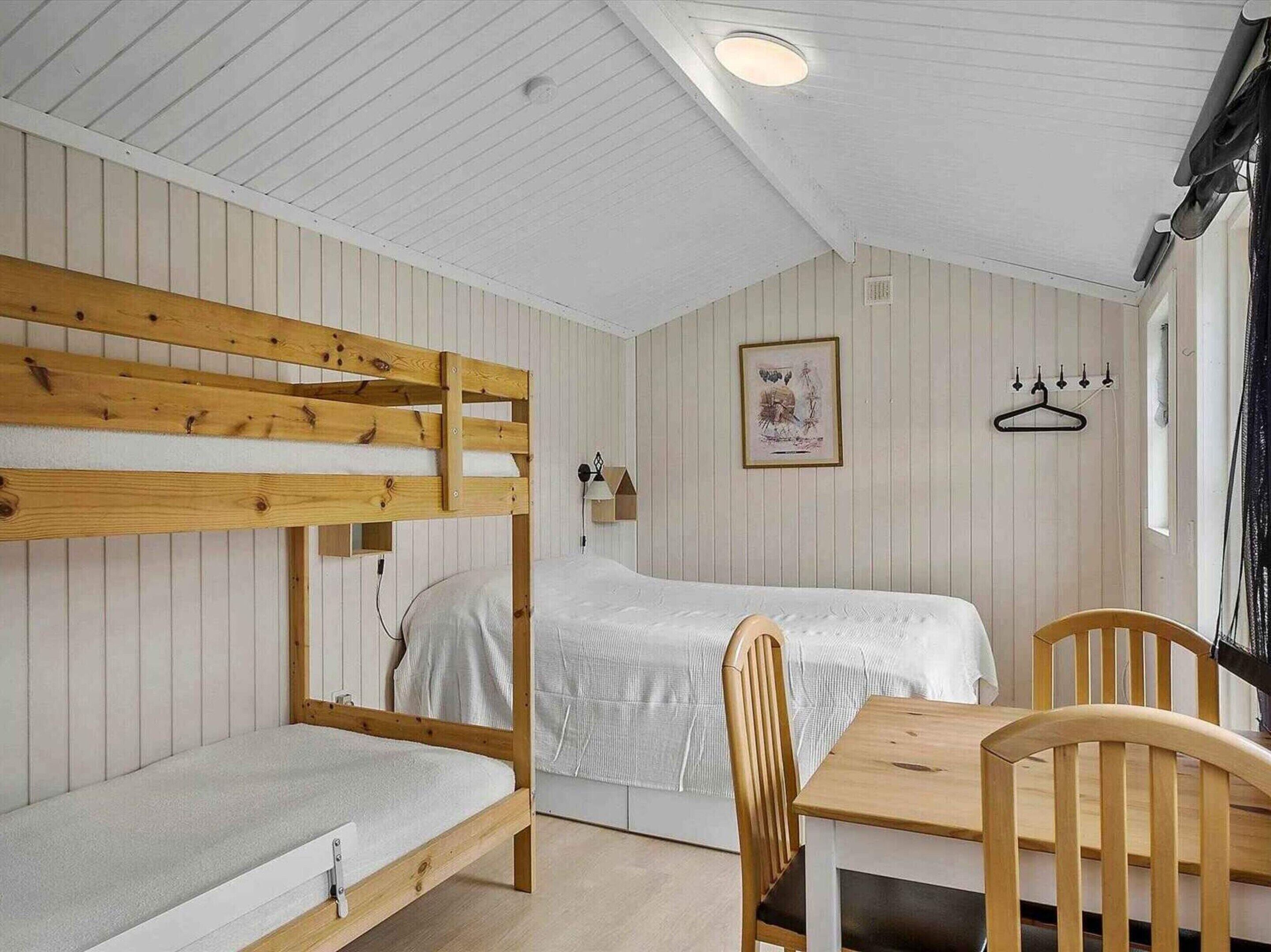 A bunk bed room with a wooden table and chairs, a framed picture on the wall, and a window with a black hanger.