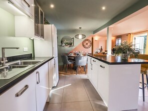 A modern kitchen with white cabinets, a black countertop, a dining area with a table and chairs, and a large wall clock. - Willow Cottage: A Cozy Escape (Rye)