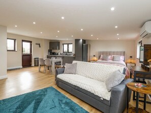 A modern, well-lit room with a grey sofa, a dining area, a kitchen, and a large bed with a headboard. - Aloe Lodge Retreat Near Heath Fm (Diss)
