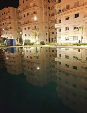 A multi-story hotel with balconies, a swimming pool with a clear reflection, and street lamps.