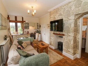 A cozy living room with a sofa, a television mounted on a stone wall, a fireplace, and a kitchen in the background. - Fox Cottage, Droitwich Spa (Droitwich)