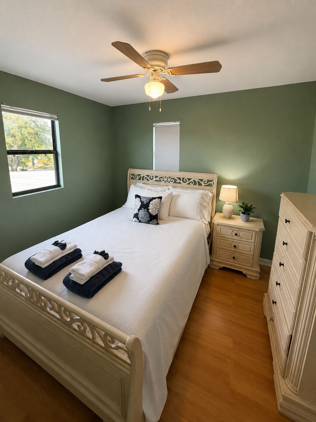 A bedroom with a bed, a nightstand, a dresser, a ceiling fan, a window, and a wall-mounted light.