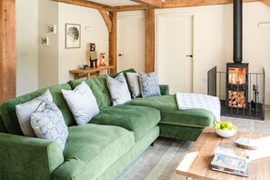 Een groene bank met gestreepte kussens, een houten bijzettafel met boeken en een schaal appels, en een houtkachel.