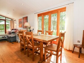 A dining area with a wooden table and chairs, large windows with curtains, and a view of greenery outside.