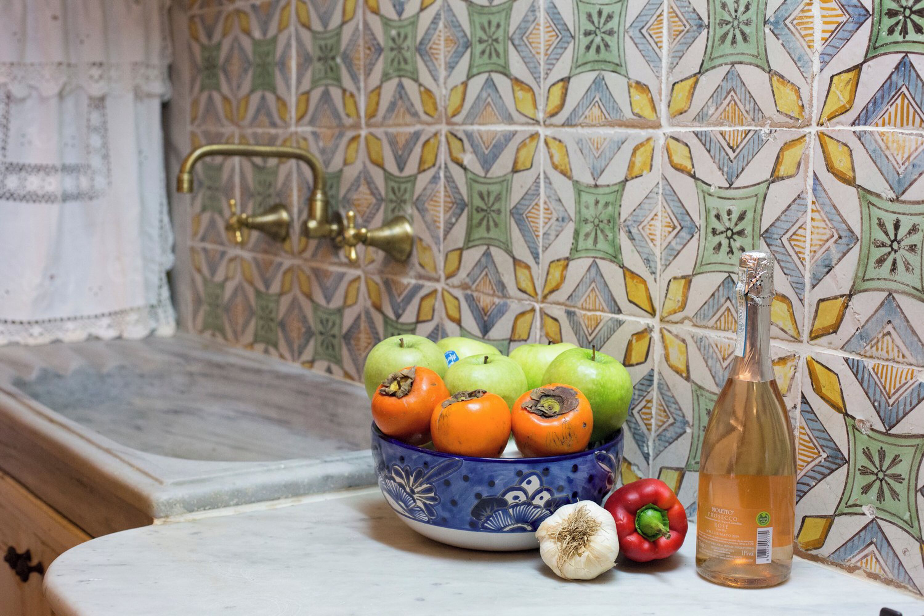 A kitchen with a patterned tile backsplash, a blue bowl with apples and tomatoes, a bottle, and a red bell pepper.