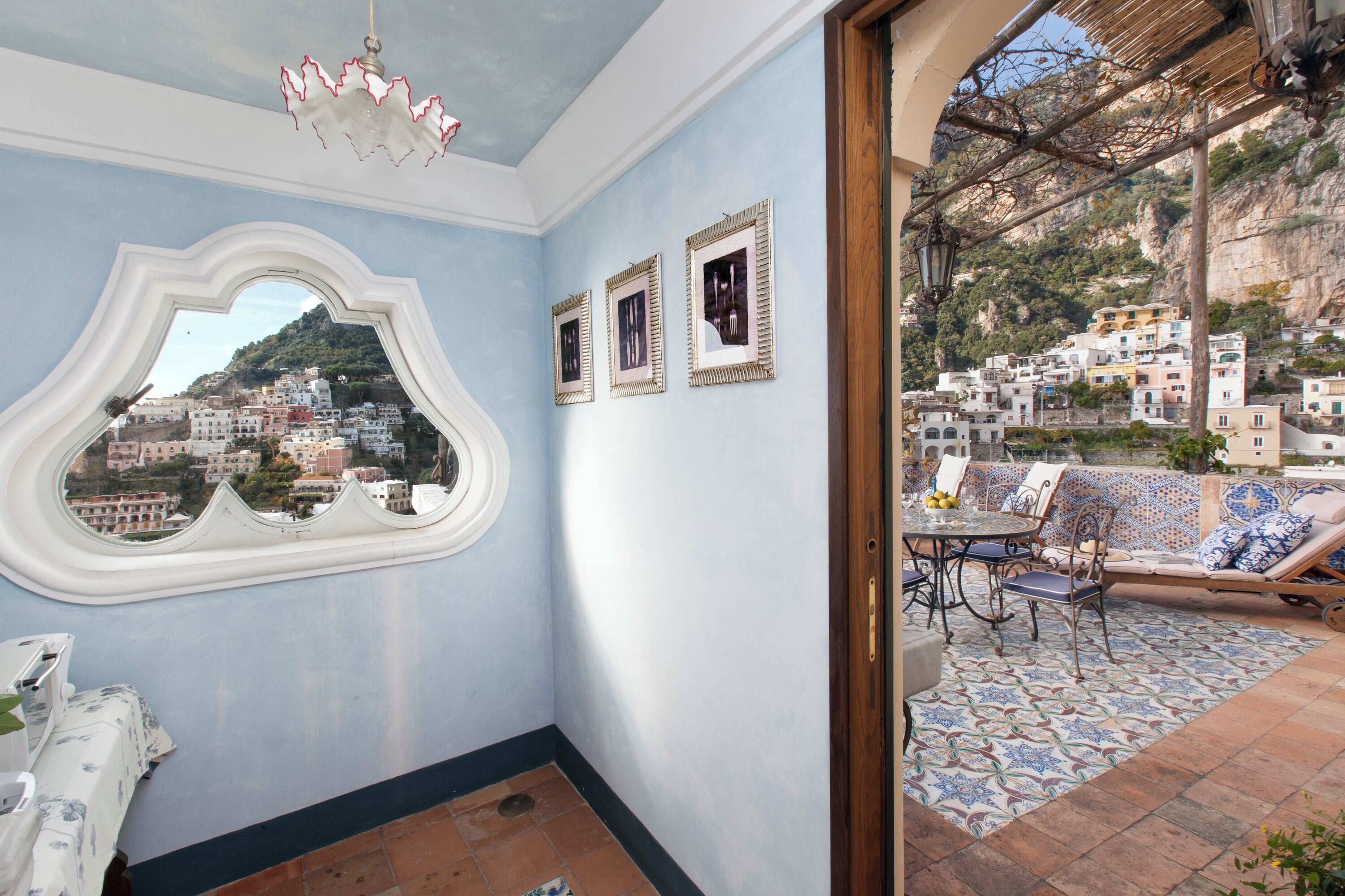 A room with a view of a coastal town, a window with a decorative frame, a dining area with a table and chairs, and a patterned floor.