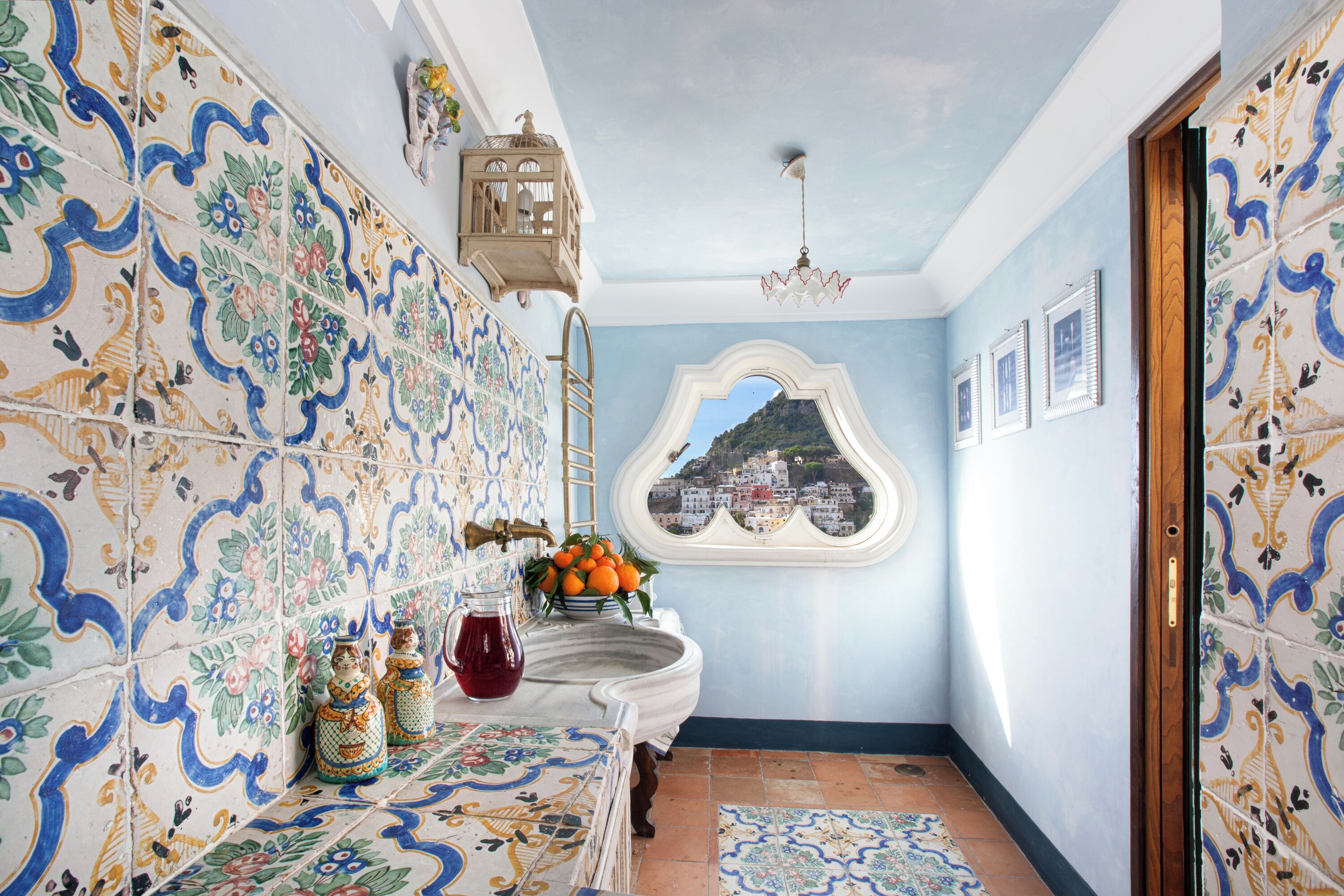 A bathroom with ornate tiles, a sink, a pitcher, and a window with a view of a hill.