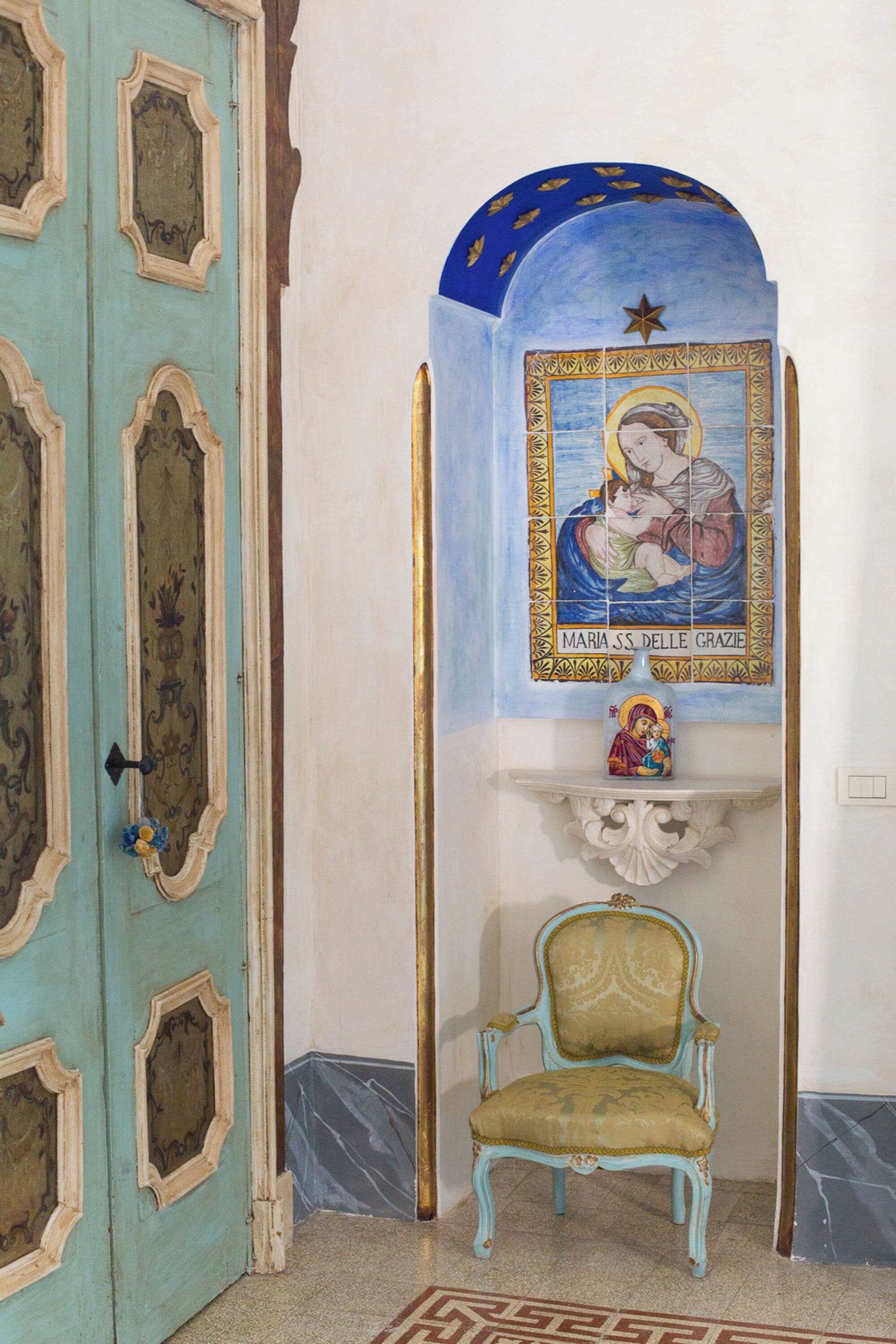 Um quarto com porta azul-turquesa, um mural dourado e azul, uma estante branca com uma estátua, e uma cadeira com moldura dourada.