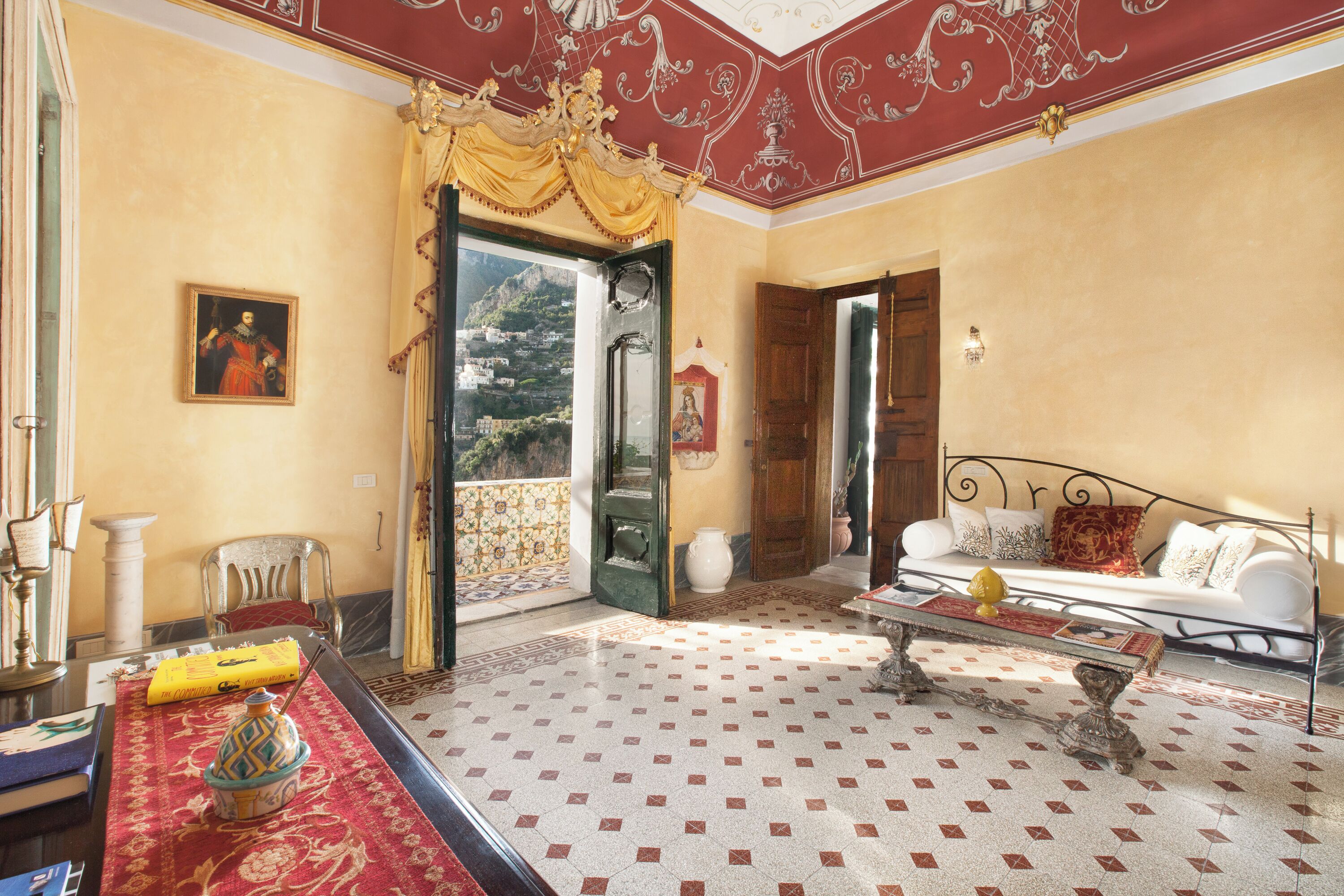 A room with a patterned floor, a sofa, a table with a vase, a chair, a painting, and a door leading to a balcony.