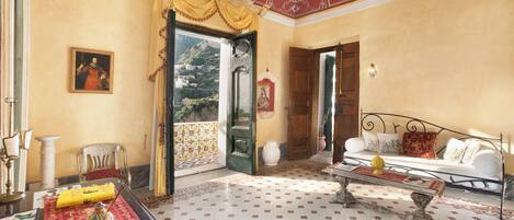 A room with a patterned floor, a sofa, a table with a vase, a chair, a painting, and a door leading to a balcony.