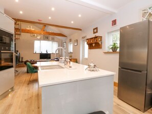 A modern kitchen with a central island, stainless steel appliances, wooden beams on the ceiling, and a view of the dining area. - Primose Cottage, Cromer, Norfolk (Cromer)