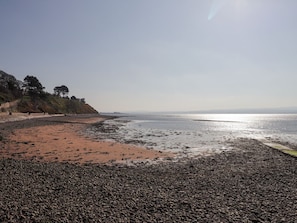 Ein kiesiger Strand mit blauem Himmel, einer fernen Küstenlinie und Bäumen an einem Hügelhang.