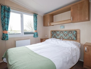 A bedroom with a bed, a window with curtains, a radiator, and wooden cabinets. - No.15 Maple Forest, White Cross Near Newquay (New Quay)
