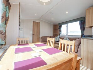 A wooden dining table with purple placemats, a wooden chair, a sofa, a window with curtains, and a kitchen area. - No.15 Maple Forest, White Cross Near Newquay (New Quay)