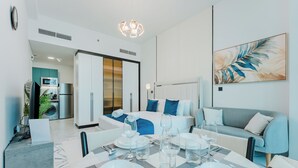 A modern hotel room with a dining area, a bed with blue bedding, a sofa, and a large abstract painting on the wall. - PVH - Opalz By Danube - Studio with Pool (Dubai)