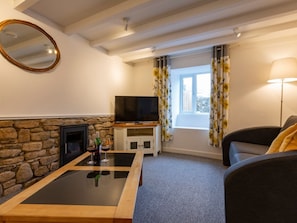 A living room with a stone fireplace, a flat-screen TV, a glass coffee table, a sofa, and a window with curtains. - Rose Cottage, St Ives (St. Ives)