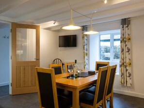 A dining area with a wooden table, four chairs, a television on the wall, and a window with curtains. - Rose Cottage, St Ives (St. Ives)